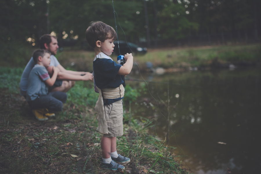 Young child fishing at the edge of a calm pond with simple rod and reel while family watches nearby.