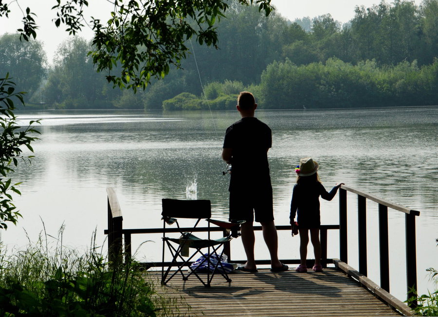 A man and a child fishing from a wooden dock by a calm lake.