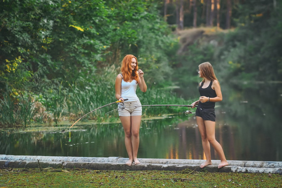 Two young women fishing by a serene lake surrounded by greenery.