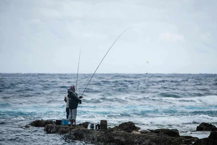 Two anglers fishing from rocky shorelines against a backdrop of ocean waves.