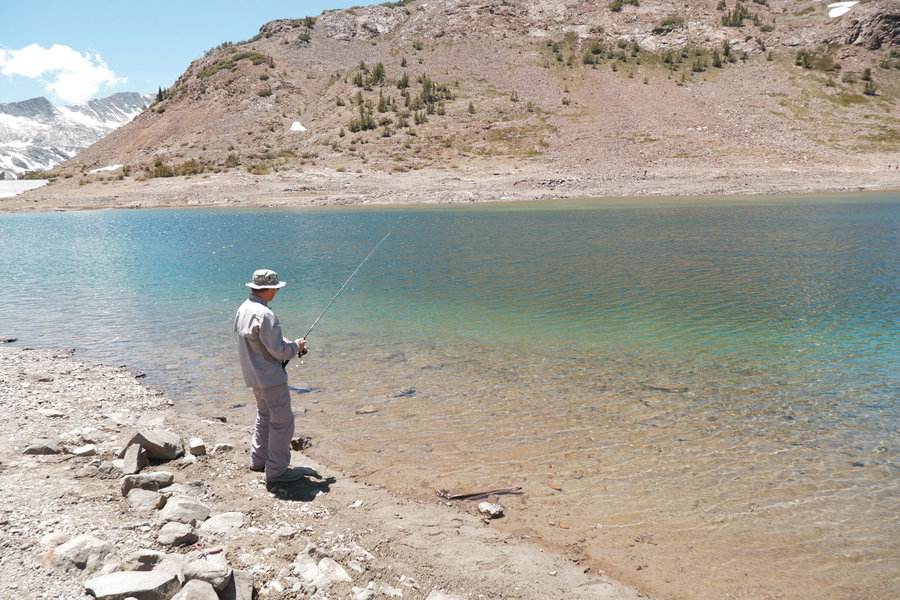 Angler fishing by a clear blue lake surrounded by mountains and rocky shore.
