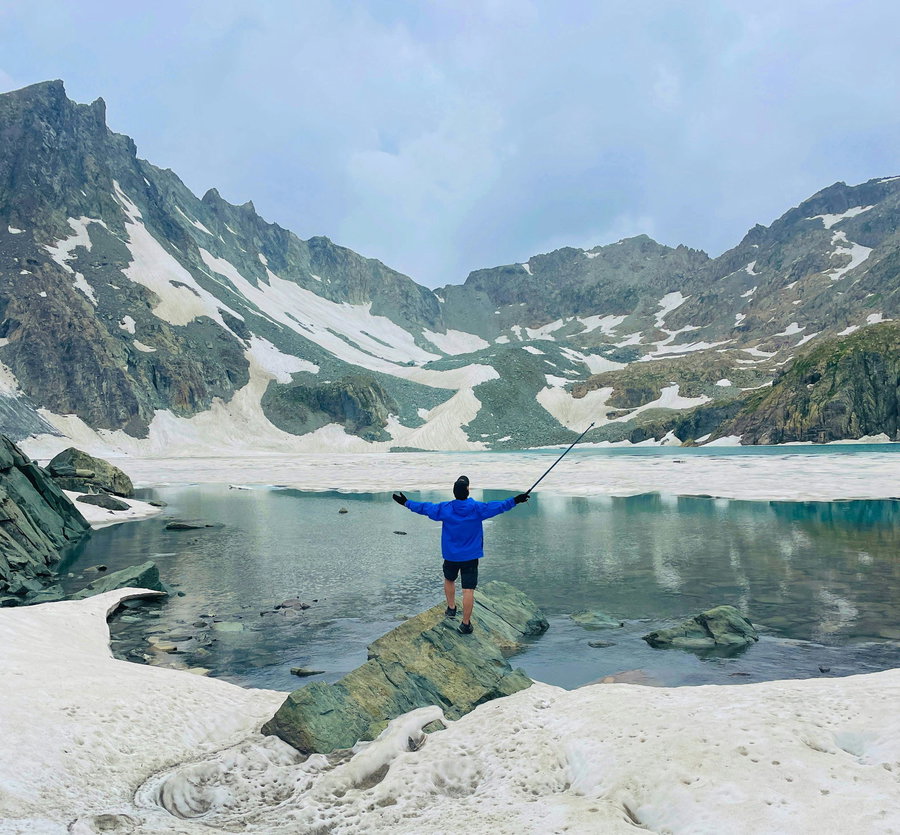 Person fishing on a rocky shore with mountains and snow in the background.