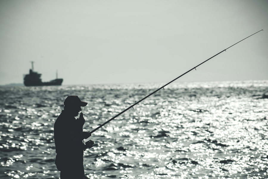 Silhouette of a person fishing by the shimmering sea with a distant ship.