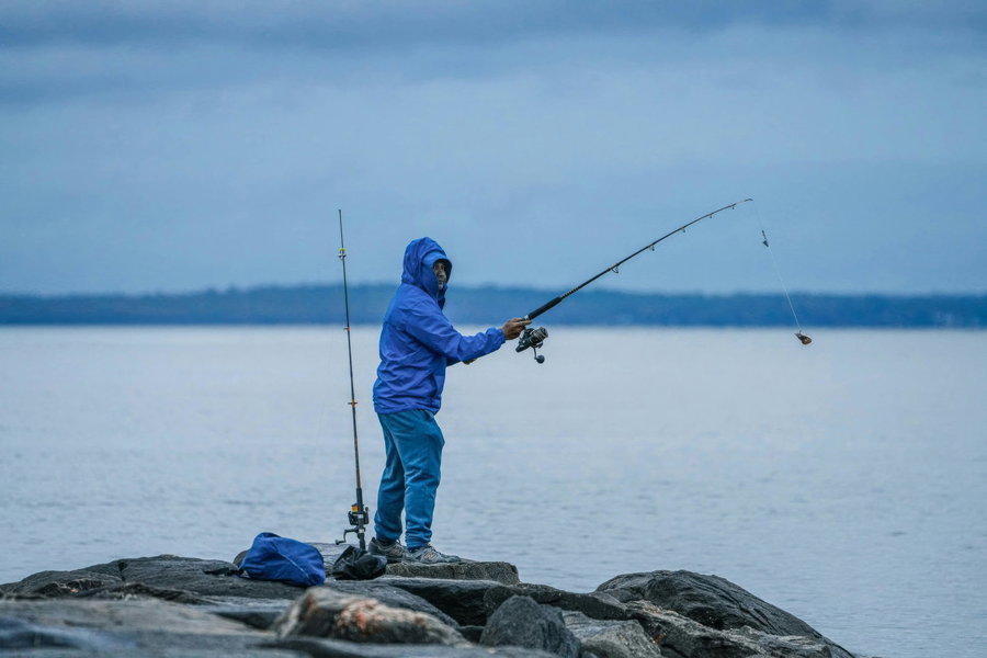 Person fishing from rocky shore, casting a line into calm water.