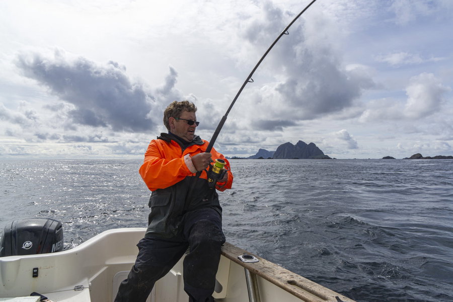 Fisherman reeling in a line on a boat with rocky islands in the background.