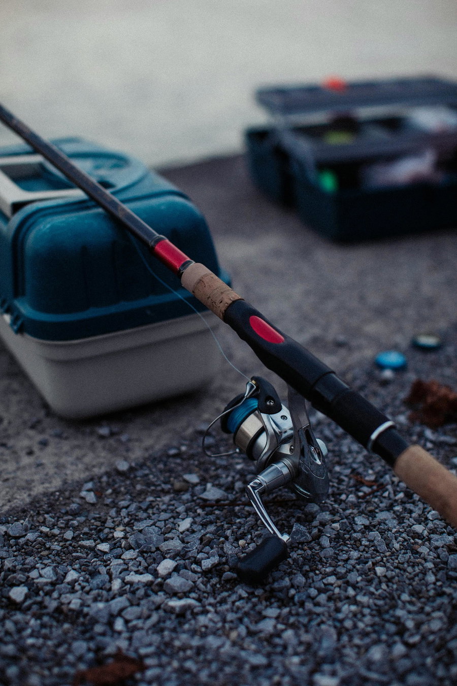 Fishing rod with reel resting on gravel near a tackle box.