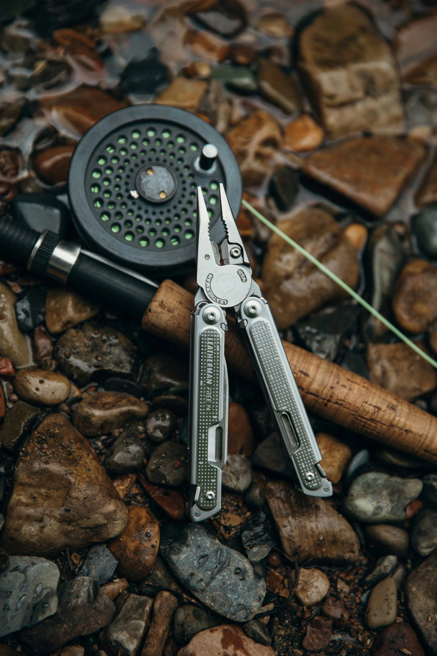 Multi-tool pliers resting on a fishing rod and reel, surrounded by wet stones.
