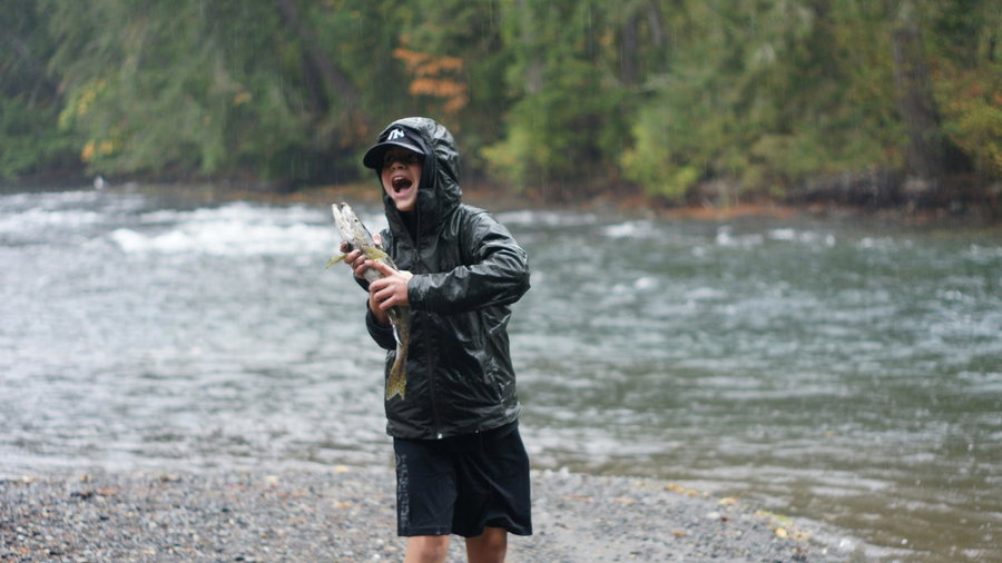 Child in rain gear joyfully holds a fish by a river.