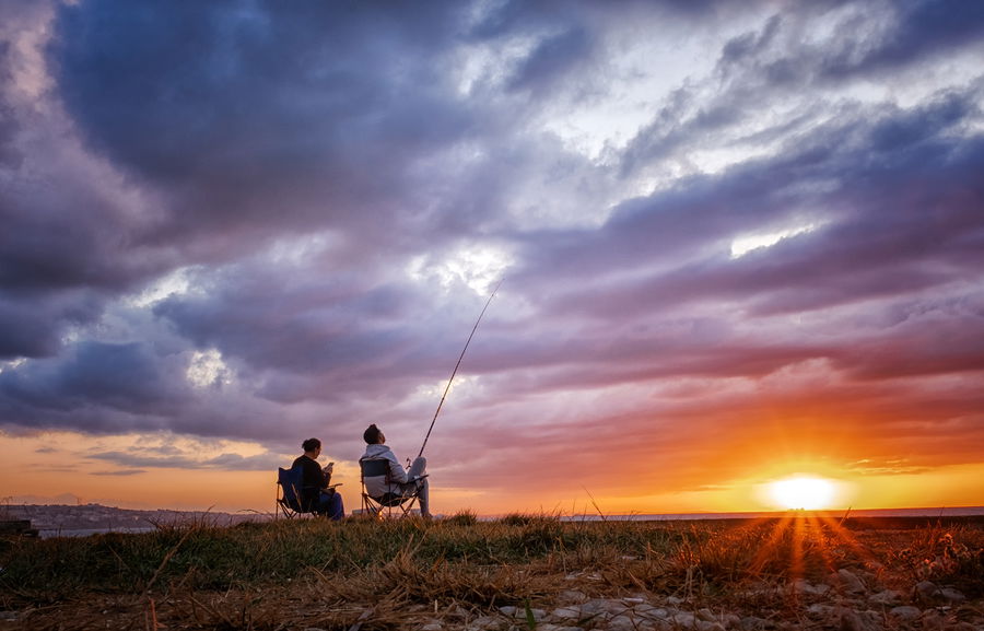 Two anglers fishing at sunset on a grassy shoreline, dramatic sky and golden light reflecting over calm water.