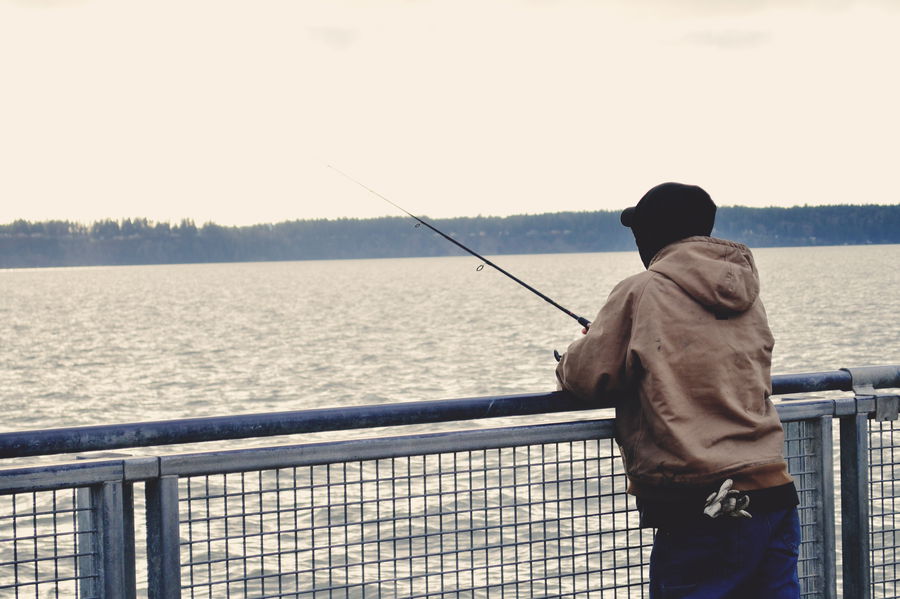 Beginner angler standing by a railing over calm water, patiently fishing and looking toward the lake horizon.